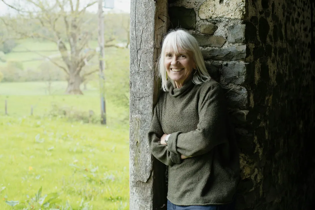 A smiling woman with white hair leaning on an old stone wall